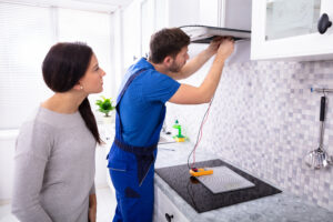 Male Repairman Checking Extractor Filter With Digital Multimeter While Woman Standing In Kitchen